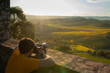 A photographer in a yellow shirt captures the sunset from an ancient wall, with a picturesque landscape in the background