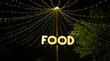 Signboard with word food made with light bulbs hung on a wooden post under light garlands located in the middle of treetops at night. Concept of food and drink zone on outdoors summer festival.