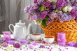 A basket of purple flowers sits on a table with a white tea set and candles