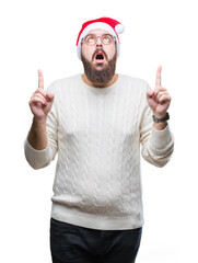 Poster - Young caucasian man wearing christmas hat and glasses over isolated background amazed and surprised looking up and pointing with fingers and raised arms.