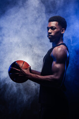 Wall Mural - Basketball player holding a ball against blue fog background. Muscular african american man looking at camera.