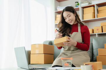 Wall Mural - Young Asian business woman smiles and uses laptop to check information on shipping box before sending it to customer. Small business entrepreneur, SME online marketing, delivery concept