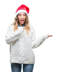 Poster - Beautiful young blonde woman wearing christmas hat over isolated background amazed and smiling to the camera while presenting with hand and pointing with finger.