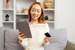 Young Asian Woman Shopping Online On Tablet At Home, Smiling As She Holds A Credit Card, Cozy And Comfortable Living Room Setting