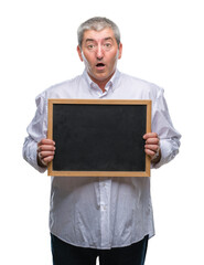 Canvas Print - Handsome senior teacher man holding blackboard over isolated background scared in shock with a surprise face, afraid and excited with fear expression