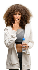 Wall Mural - African american fitness woman holding water bottle cover mouth with hand shocked with shame for mistake, expression of fear, scared in silence, secret concept