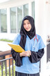 Asian Female doctor in hijab portrait of cheerful happy holding a clipboard in medical blue shirt and glasses with stethoscope on modern clinic office background.