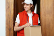 Friendly Delivery Man With Package Standing At Door, Smiling Service Worker In Red Vest And White Cap. Express Parcel Delivery, E-Commerce, Customer Satisfaction