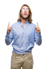 Poster - Young handsome man with long hair wearing glasses over isolated background amazed and surprised looking up and pointing with fingers and raised arms.