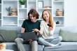 Happy Couple Shopping Online At Home. Young man and woman sitting on a couch, smiling as they make a purchase with a credit card and laptop in a cozy living room.