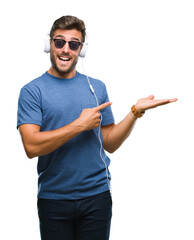 Poster - Young handsome man wearing headphones listening to music over isolated background amazed and smiling to the camera while presenting with hand and pointing with finger.