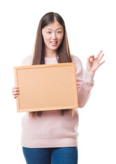 Poster - Young Chinese woman over isolated background holding corkboard doing ok sign with fingers, excellent symbol