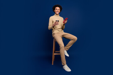 Poster - Full size photo of nice young man sit chair paper cup device wear trendy beige outfit isolated on dark blue color background