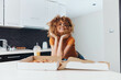Young woman with curly hair sitting on kitchen counter with pizza box in front of her, enjoying a tasty meal