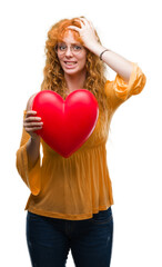 Wall Mural - Young redhead woman in love holding red heart stressed with hand on head, shocked with shame and surprise face, angry and frustrated. Fear and upset for mistake.