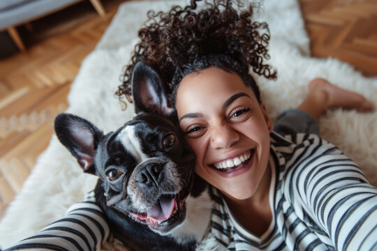 A joyful African American woman is taking a selfie with her cheerful pet, who has a funny expression on its face, and they are lying on the living room floor and making funny faces. Emotional