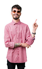 Canvas Print - Young handsome man wearing sunglasses over isolated background with a big smile on face, pointing with hand and finger to the side looking at the camera.