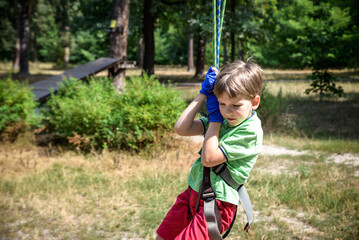 Strong excited young boy playing outdoors in rope park. Caucasian child dressed in casual clothes and sneakers at warm sunny day. Active leisure time with children concept