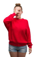 Wall Mural - Young blonde woman wearing bun and red sweater peeking in shock covering face and eyes with hand, looking through fingers with embarrassed expression.