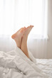 Feet crossed up in air, covered by blanket in neutral white bedroom, soft natural sunlight, lifestyle. Good sleep concept, healthy lifestyle, self care, rest and leisure pastime