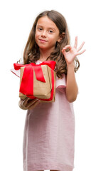 Poster - Brunette hispanic girl holding a gift doing ok sign with fingers, excellent symbol