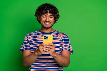 Sticker - Portrait of young curly haired guy in striped t shirt happy using smartphone eshopping features isolated on green color background