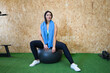 Young and beautiful brunette woman resting while sitting on a gym ball. She has a towel around her neck and is dressed in a top and leggings. Concept of health and sport.