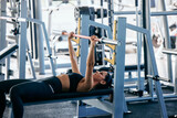 Fototapeta Pomosty - A strong girl doing exercise for her chest, holding a barbell, laying on the bench, at the gym.