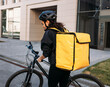 Back view of a female courier standing on a street with a bicycle. Rear view of a woman working on delivery service wearing a helmet and yellow backpack.