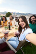 © CarlosBarquero - Vertical. Group of diverse friends having fun at rooftop party. Beautiful woman taking selfie at barbecue dinner time. Smiling people eating and drinking on outdoor terrace. Positive friendship