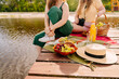 Two happy women enjoying a picnic by the water with food, tableware, and plants