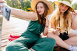 Two girls in green fashion hats happily taking a selfie in the sunlight on grass