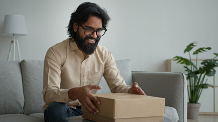 Happy male customer client consumer Arabian indian man guy in glasses open cardboard box at home unpack online shopping order purchase wrapped package get delivery present gift unpacking carton parcel