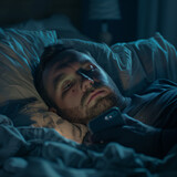 Fototapeta  - Hombre acostado en la cama sin poder dormir en medio de la noche mirando su teléfono con todas las luces apagadas