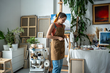 Female artist tying apron before starting work at artwork project. Happy exited woman painter getting ready for drawing process. Creative joyful girl among art paintings in home art studio.