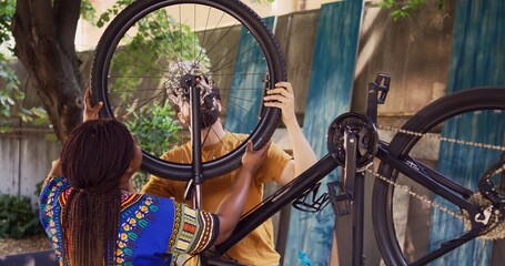 Dedicated boyfriend and girlfriend doing annual maintenance on bike tires for safe cycling in home yard. Youthful multiracial pair successfully mounting wheel on modern bicycle frame.