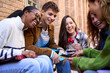 © CarlosBarquero - Group of young people students sitting in circle having fun using cell phones together outdoor. Cheerful friends looking at each other laughing enjoying funny videos. Addiction to technology of gen z