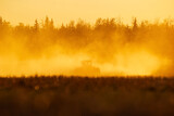 Fototapeta Maki - Silhouette of a tractor seeding in dust in the field in the orange sunset.