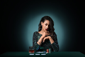 Wall Mural - Beautiful young African-American woman sitting at poker table with playing cards, chips and glass of whiskey on dark green background