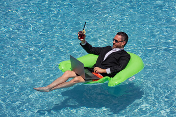 Successful business man floating in suit in pool water. Summer vacations and travel concept. Funny business man drink summer cocktail and using laptop in swimming pool. Summer business concept.