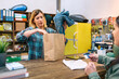 Blonde woman cashier putting jeans into a kraft paper bag while female customer holding credit card to pay purchase in local store. Female shop assistant delivering purchased clothing to client.