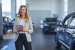 Cheerful young woman with clipboard smiling and looking at camera while working in modern car dealership. Shot of a woman using her digital tablet in a car dealership