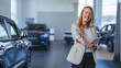 Professional young female dealer offering handshake, smiling and looking at camera friendly while standing near modern automobile in car showroom. Car dealer woman.