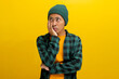 Bored young Asian man, in a beanie hat and casual outfit, rests his hand on his cheek, contemplating what to do, feeling bored, fatigued, and possibly a bit depressed. Isolated on yellow background.