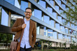 © Moon Safari - Portrait of confident manager with shoulder bag and hand in pocket standing against building in city