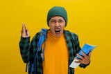 Fototapeta  - Unhappy young Asian student, dressed in a beanie hat and casual shirt and carrying a backpack, is seen expressing dissatisfaction and complaining about something while holding books, yellow background