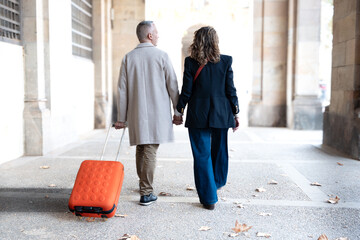 Smiling middle-aged Caucasian tourist couple walking hand-in-hand carefree with their luggage looking at each other. Happy husband and wife enjoying their outdoor vacation on a sunny winter day