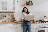 Fototapeta Do pokoju - Young woman multitasking in cozy kitchen, working remotely on laptop and talking on the phone, surrounded by stylish kitchen decor. Concept of balancing home life and work, and remote work setups