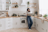 Fototapeta Do pokoju - Confident young woman freelancer successfully balancing remote work and home chores, talk on the phone call and work on laptop in a well-organized, stylish kitchen. Concept of lifestyle management
