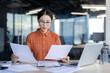 © Liubomir - A serious young professional woman in a stylish orange shirt reviews paperwork intently, surrounded by laptops in a well-lit office setting. Her focus and determination are evident.